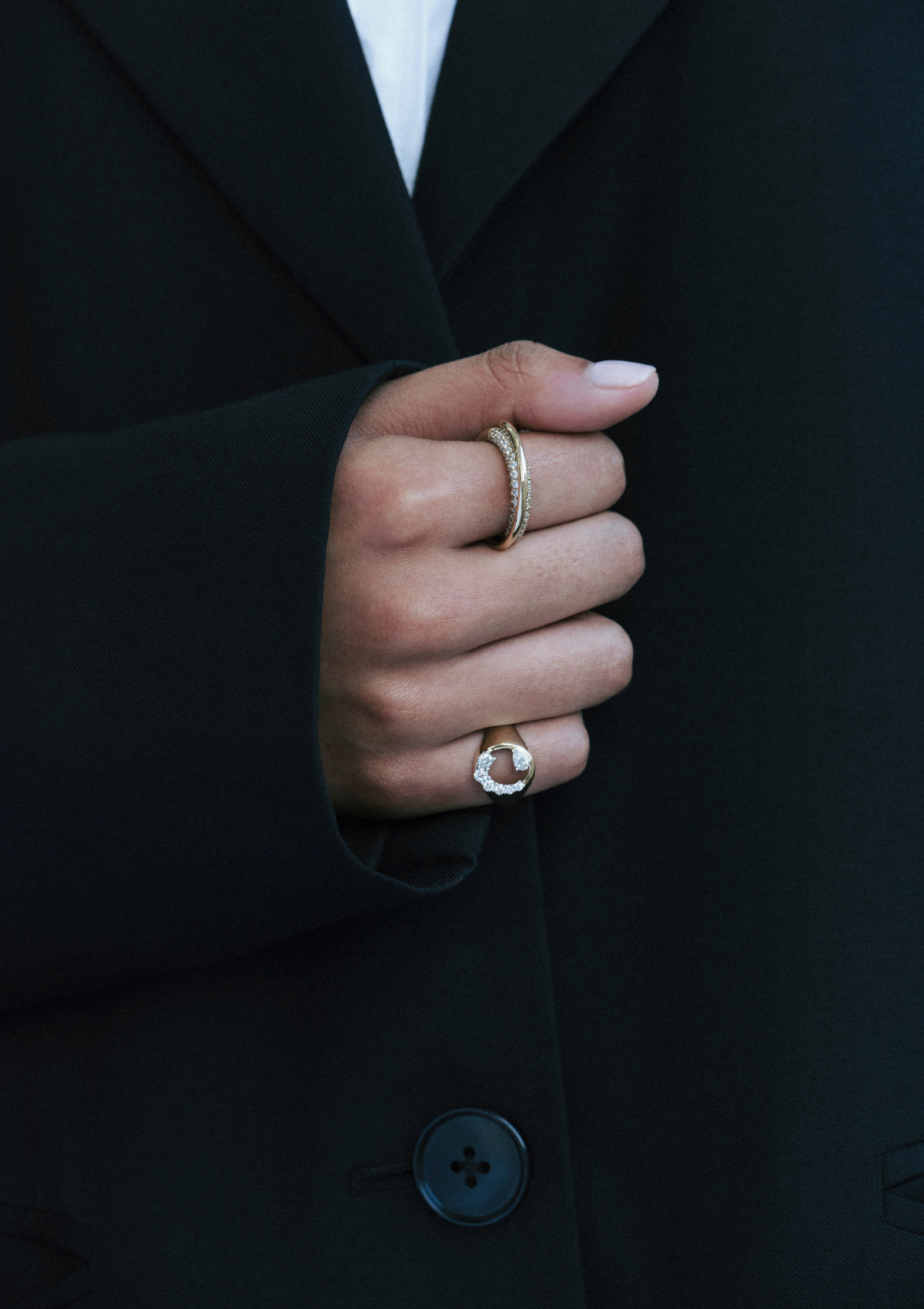 Hand wearing two rings with a black coat in the background. Pinky finger has a ring with diamonds and negative space through the middle of the ring