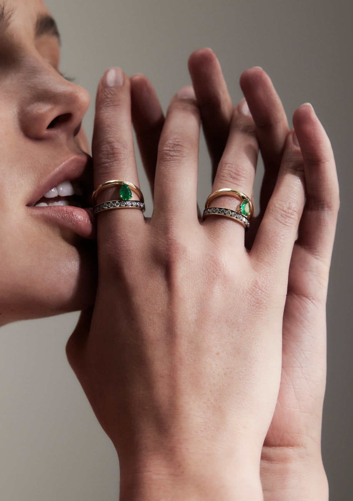 A close-up of a woman's hand showcasing a gold ring with a pear-shaped emerald, a plain asymmetric band and second band below with smaller diamonds.