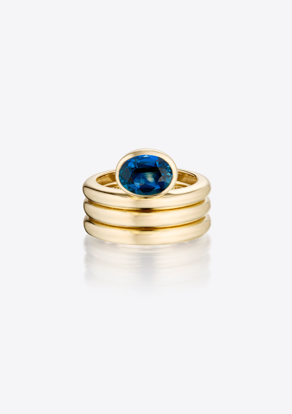 Sculptural, gold ring with a triple ribbed band and an oval blue sapphire on a grey background