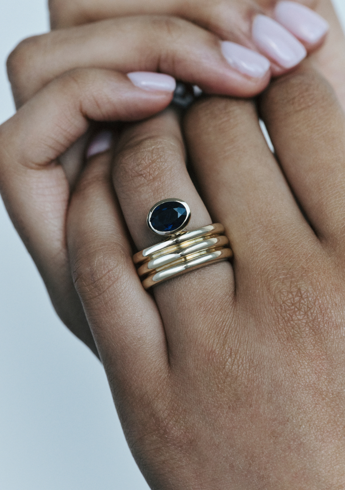 Close-up of a hand wearing a sculptural gold ring with a blue oval sapphire on a neutral background