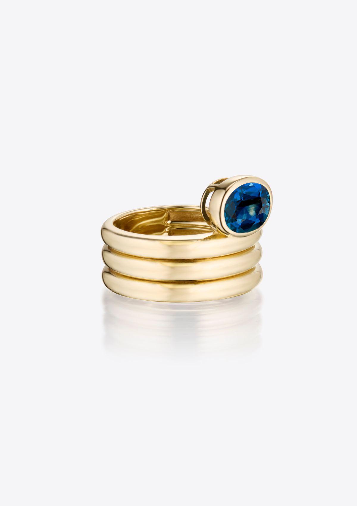 Gold ring with a sculptural, triple ribbed band with a oval blue sapphire sitting above the band. against a grey background