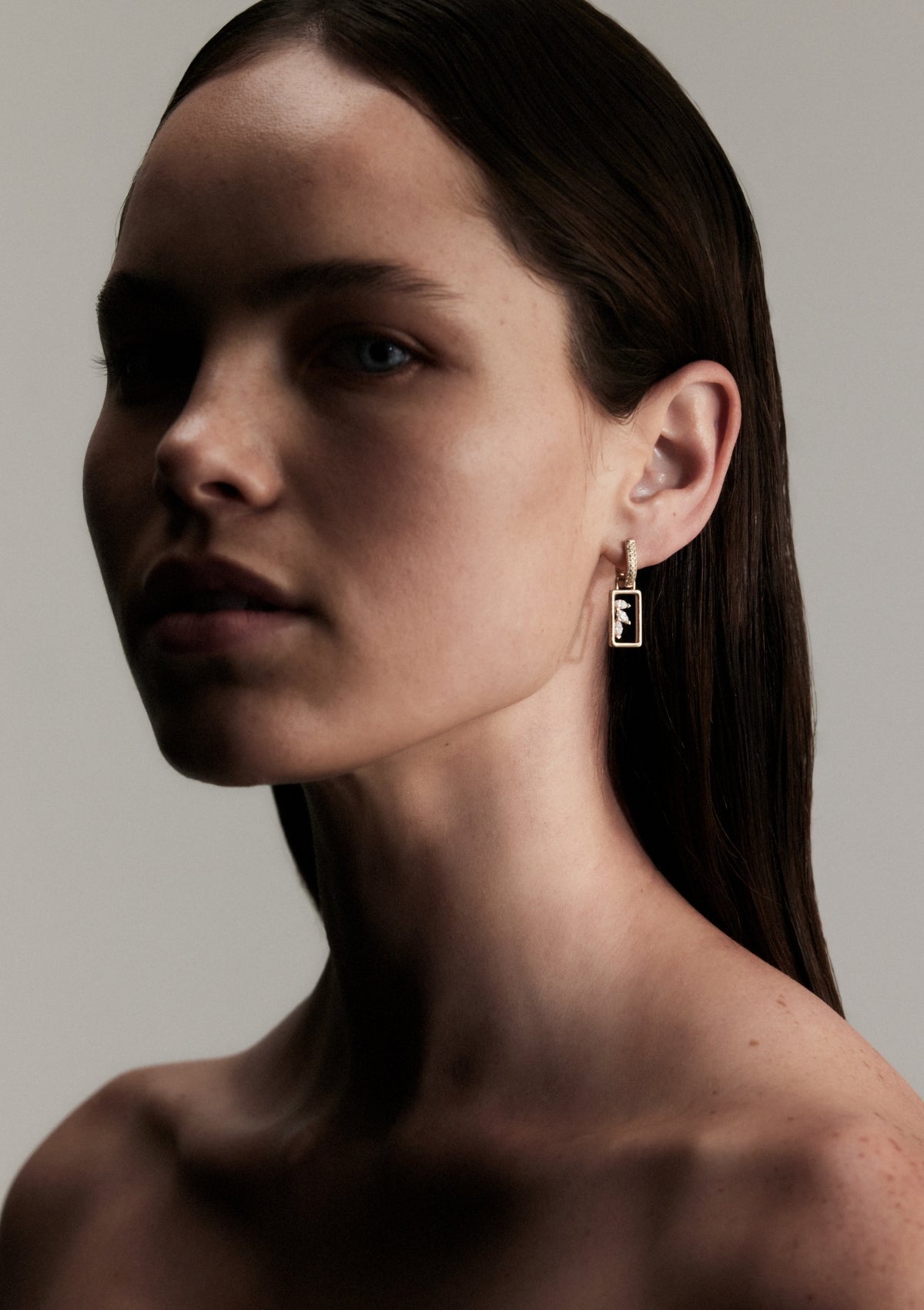 A woman wearing a pair of rectangle pendant earrings with marquise diamonds and pave huggies. against a neutral background
