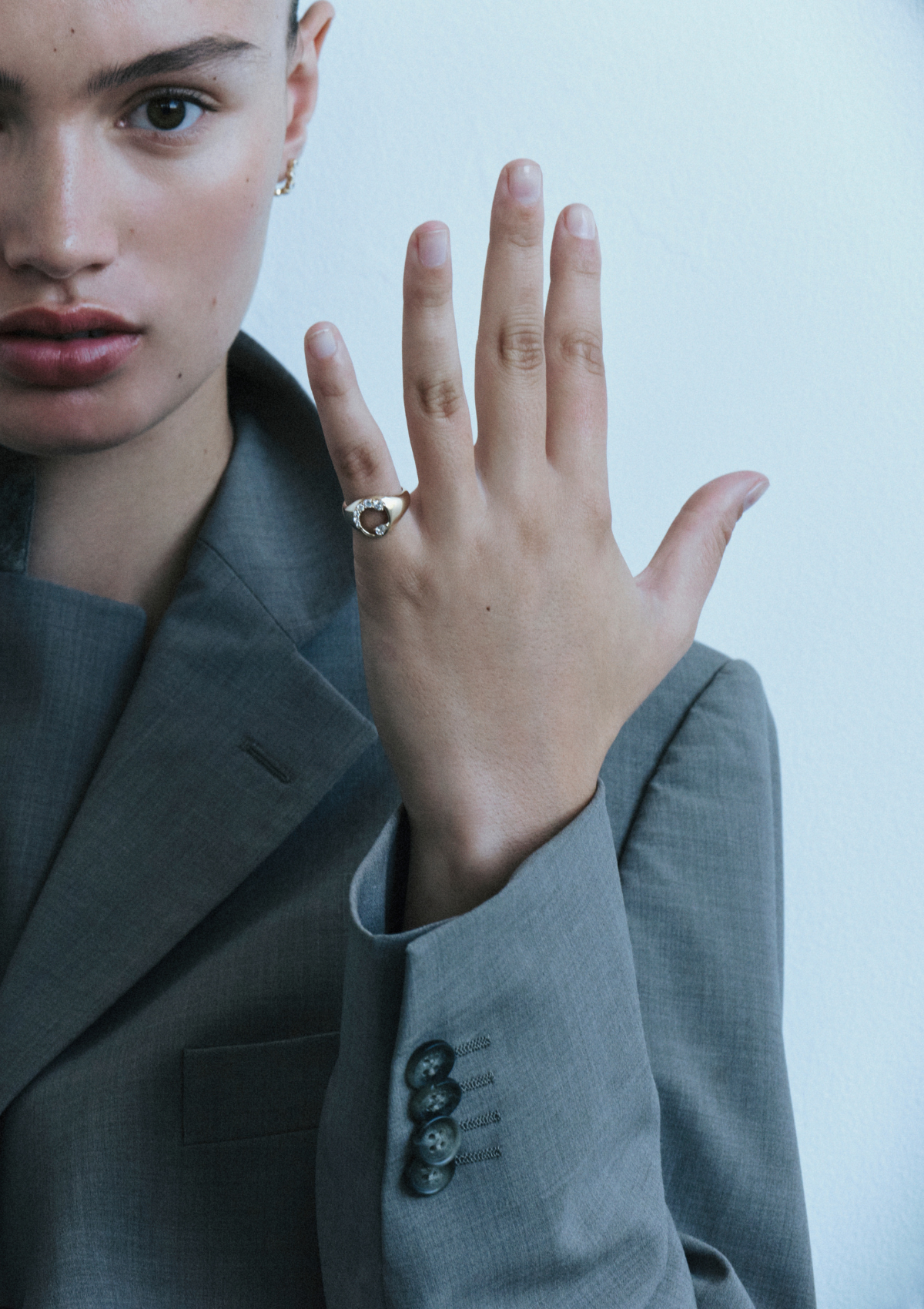 Person wearing a gray suit with a hand raised, showing a diamond ring on pinky finger against a plain background