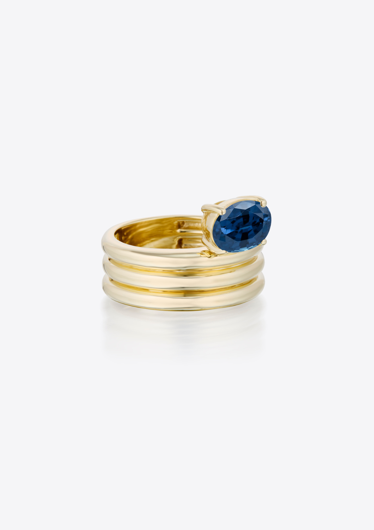Gold ring with a sculptural ribbed band and an oval blue sapphire on a grey background