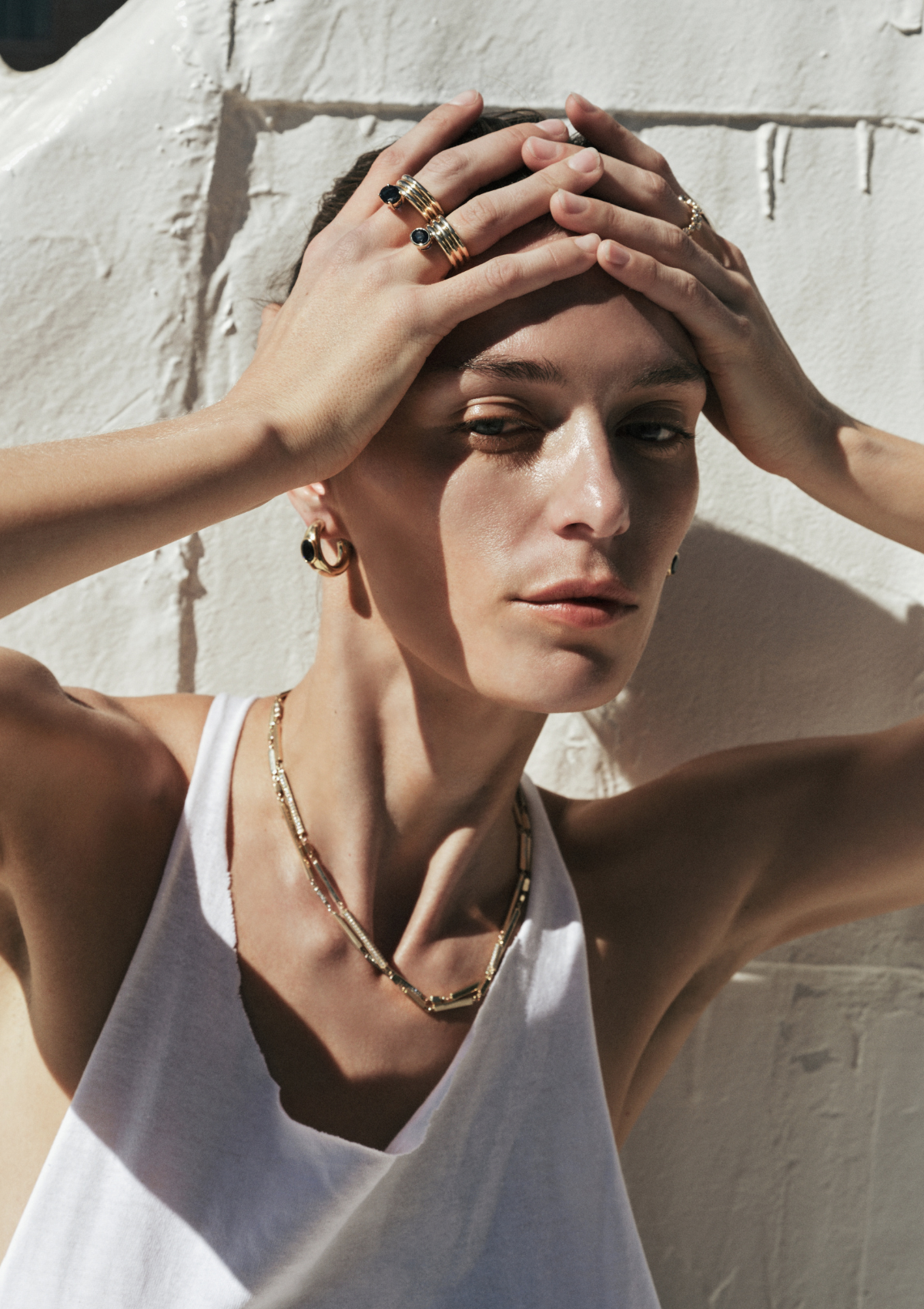 A woman wearing gold jewelry, including a layered gold chain necklace, hoop earrings, and multiple gold rings with blue sapphires, posing with her hands on her head against a textured white wall.