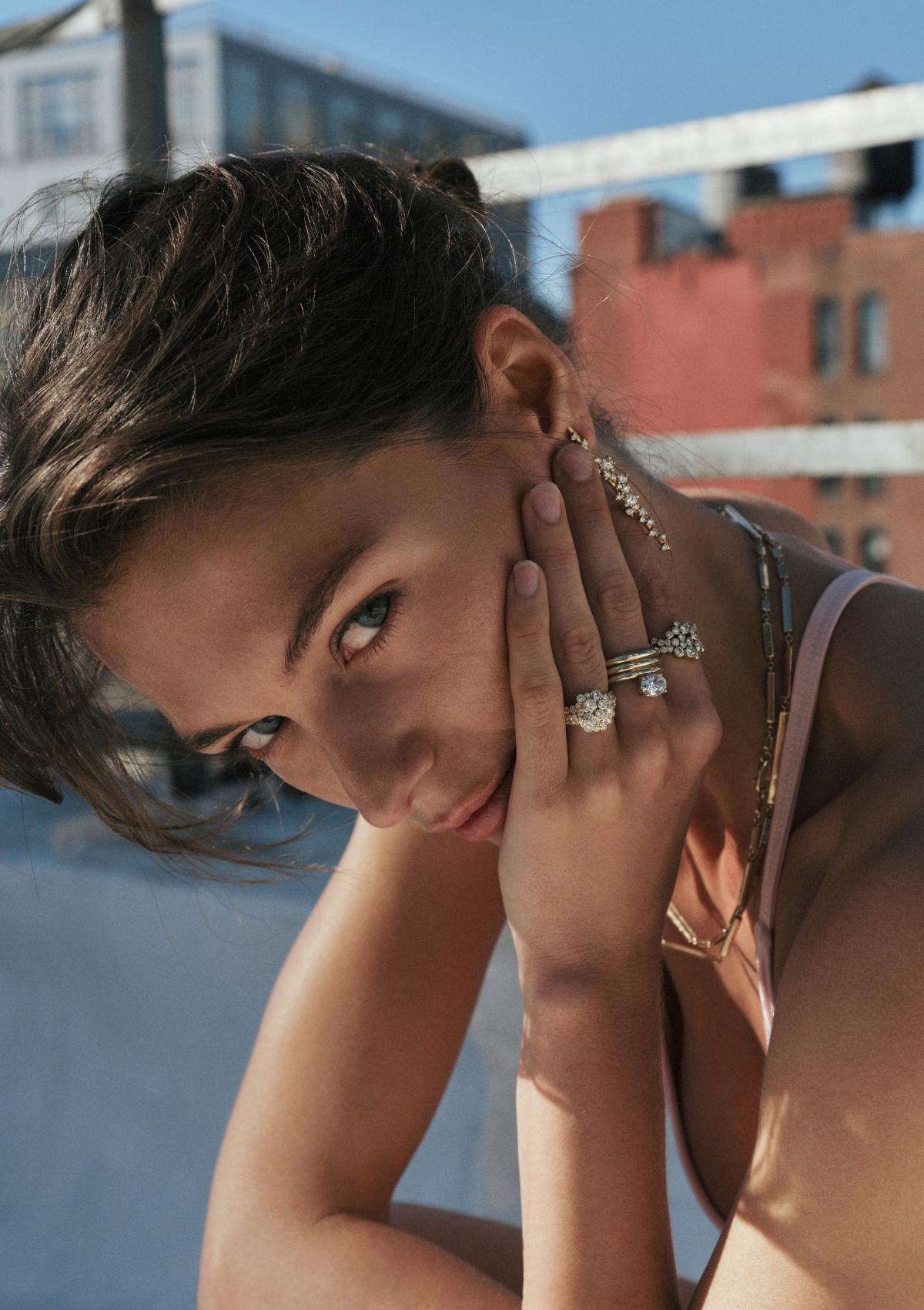 A close-up lifestyle shot of a woman with her hand resting against her face, showcasing a stack of gold rings with diamonds and a long gold diamond earring. An urban background with buildings and blue sky is visible.