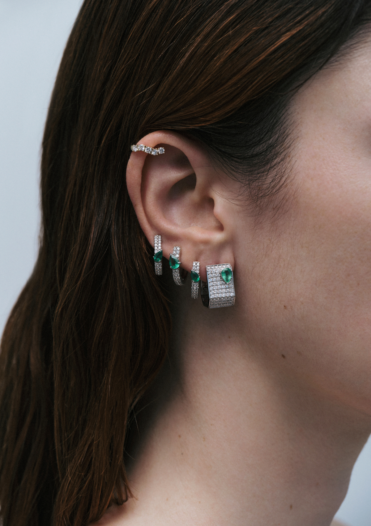 Profile view of a woman wearing asymmetrical emerald and pave diamond hoop earrings with a neutral background