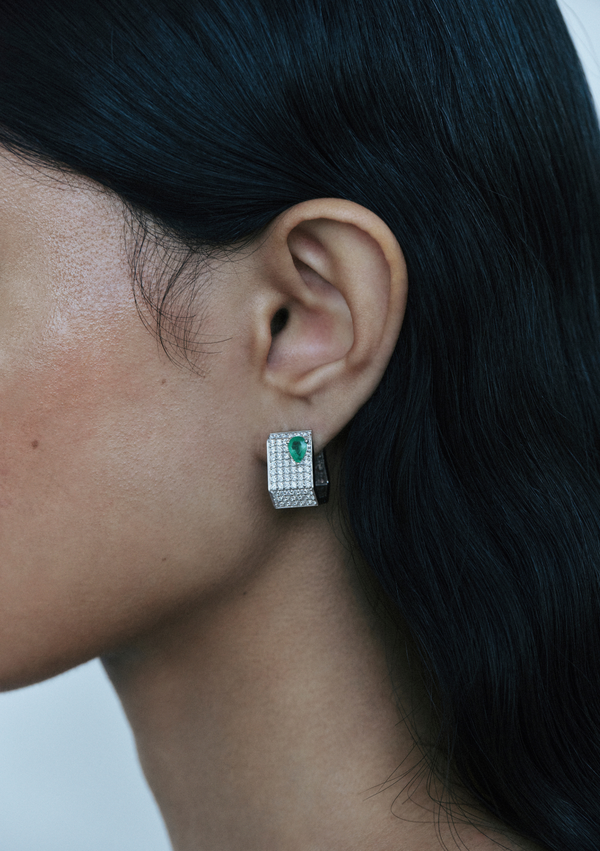 Close-up of an ear wearing a geometric shaped pave diamond earring with a pear emerald.