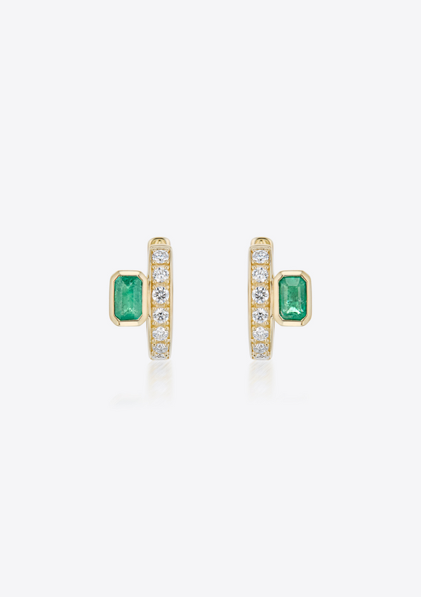 Front view of a pair of gold diamond huggies with emeralds placed off-centre on a grey background