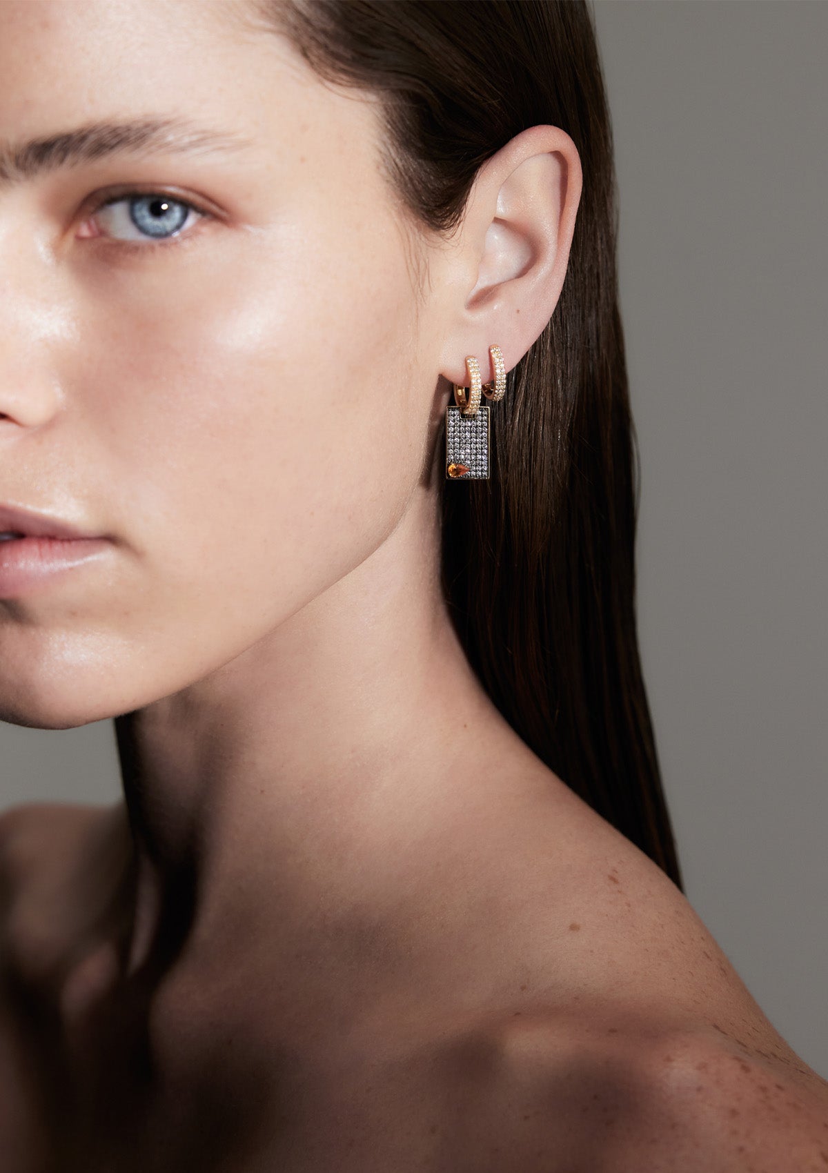 A woman's face and ear wearing a pave diamond earring with a pear-shaped orange stone and smaller diamonds around it, set against a neutral background.