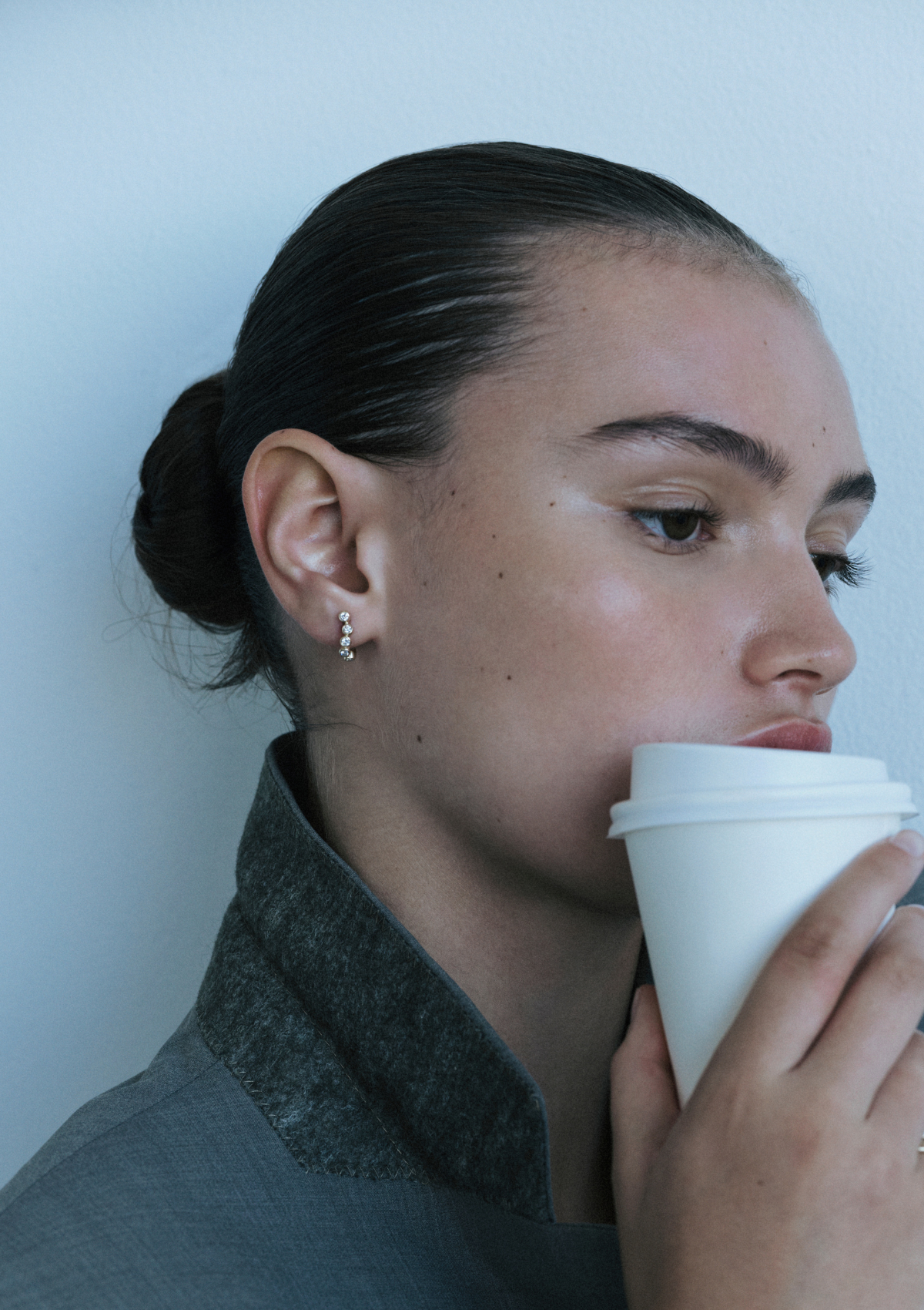 Woman drinking from a white cup wearing Gold earrings with small bezel-set diamonds shaped in a curve against a plain background.