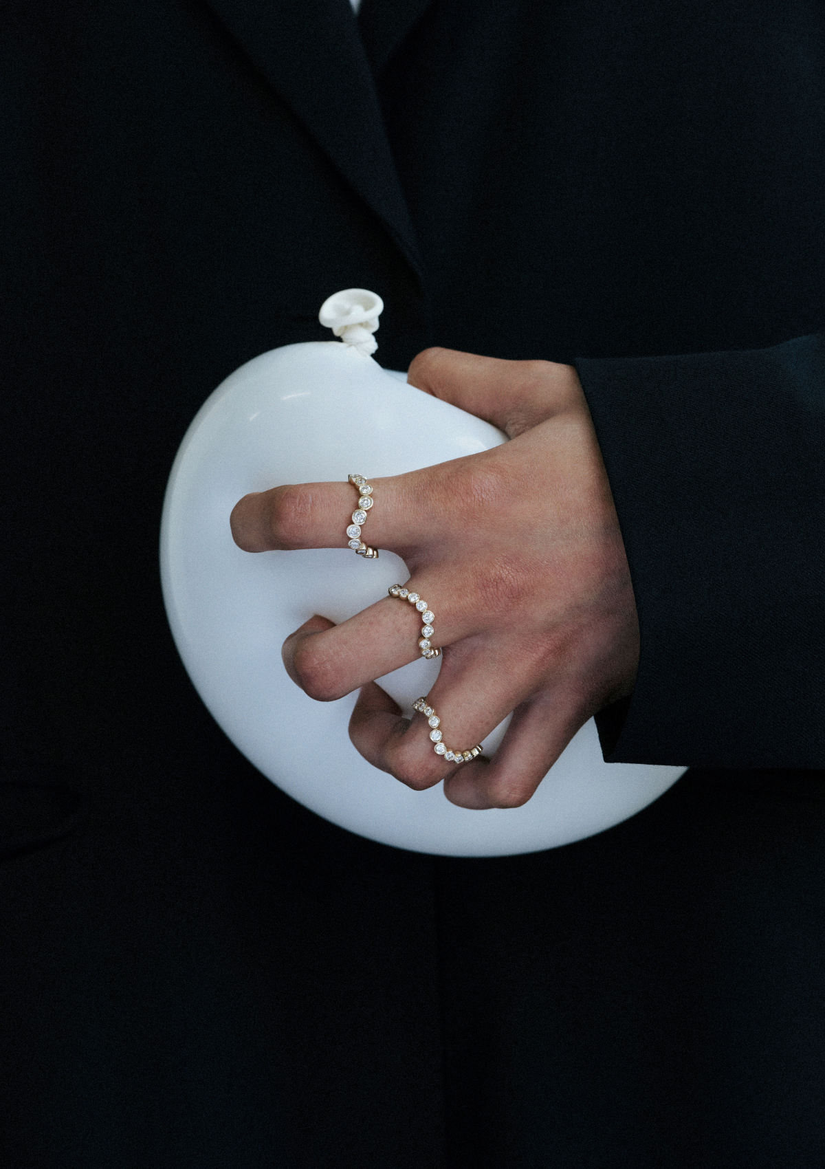 Hand holding a white balloon with curved diamond rings on 3 fingers on a black background