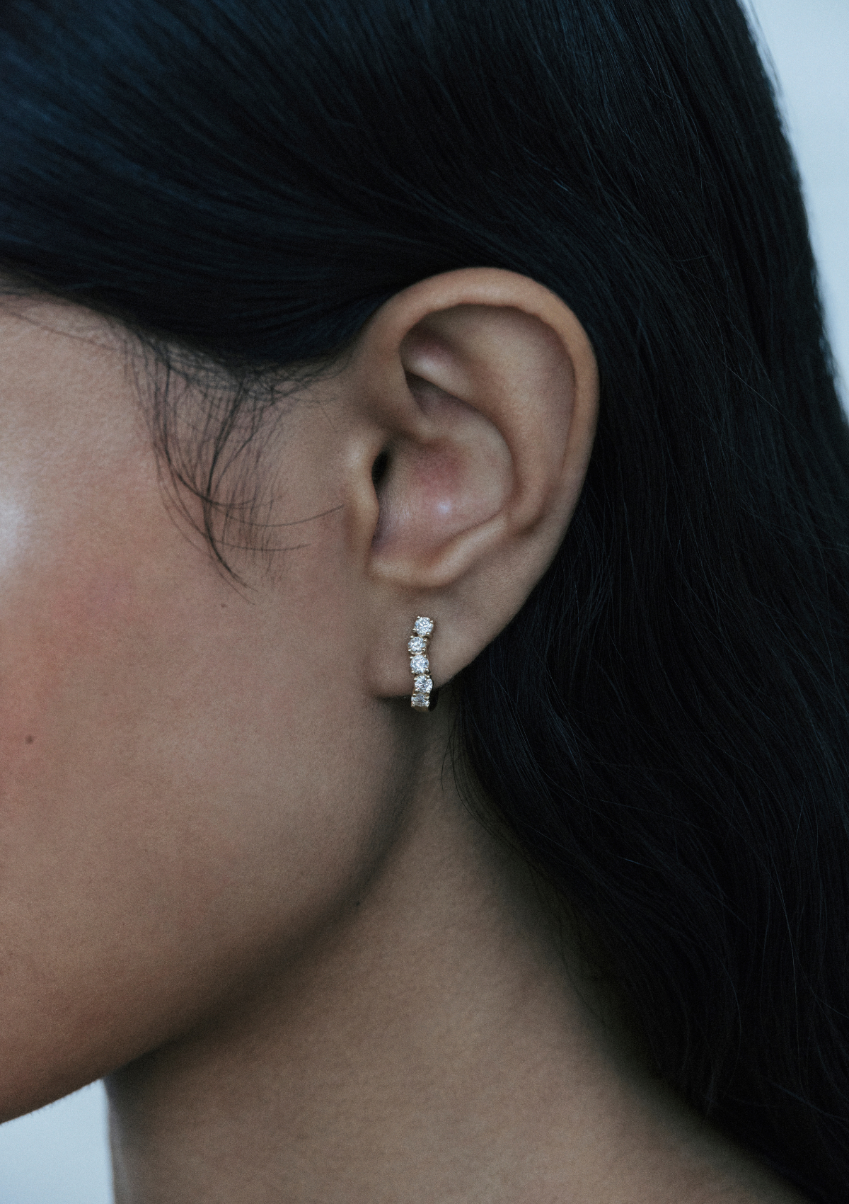 Close-up of an ear wearing a small diamond sculptural curved huggie earring with dark long hair