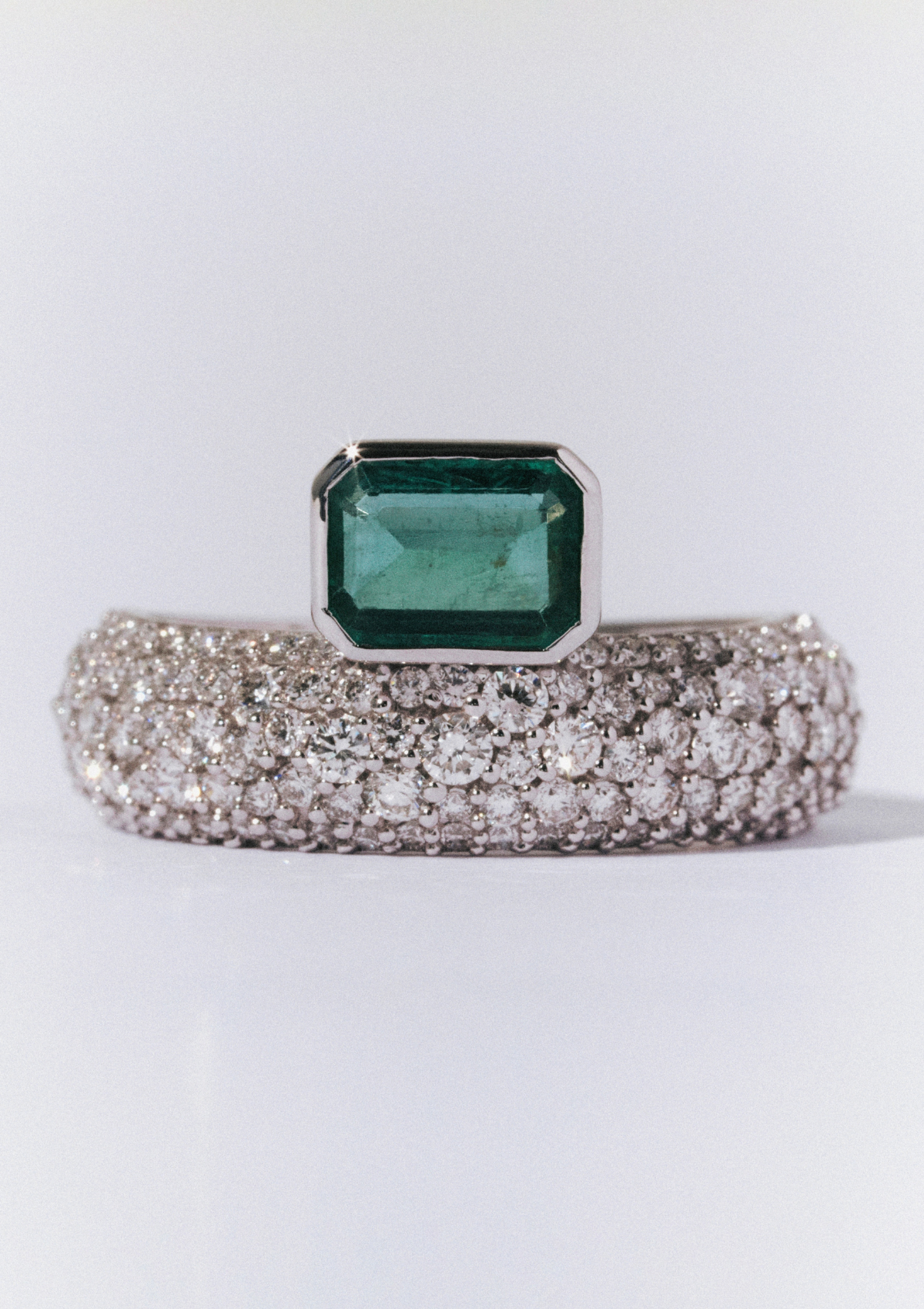 close up of white gold ring with pave diamonds and an emerald sitting on top. and a yellow gold band below. on a grey background
